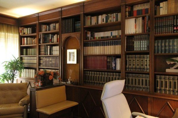 Una biblioteca domestica con ampie librerie in legno colme di libri, una sedia da scrivania e una seduta vicino a una finestra luminosa.