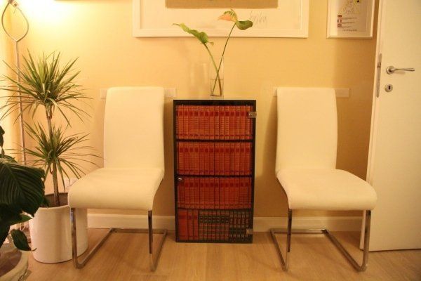 Ai lati di una piccola libreria piena di libri con il dorso rosso, accanto a una pianta in vaso, ci sono due sedie bianche.