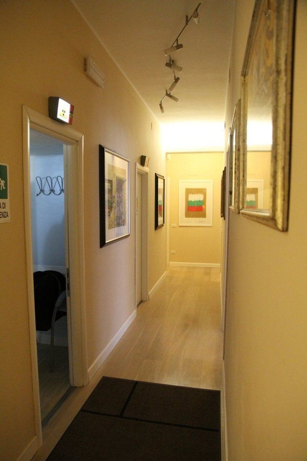 Un luminoso corridoio interno con pareti beige, quadri incorniciati, pavimento in legno chiaro e illuminazione a incasso.