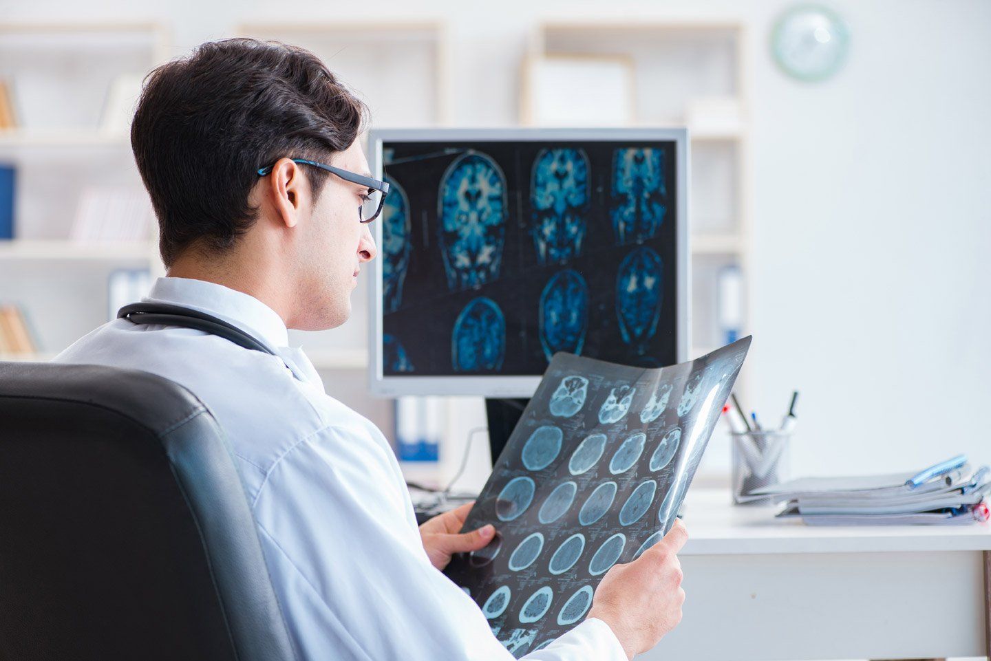 Un medico in camice bianco esamina una risonanza magnetica mentre è seduto a una scrivania con un monitor che mostra le immagini della scansione cerebrale.