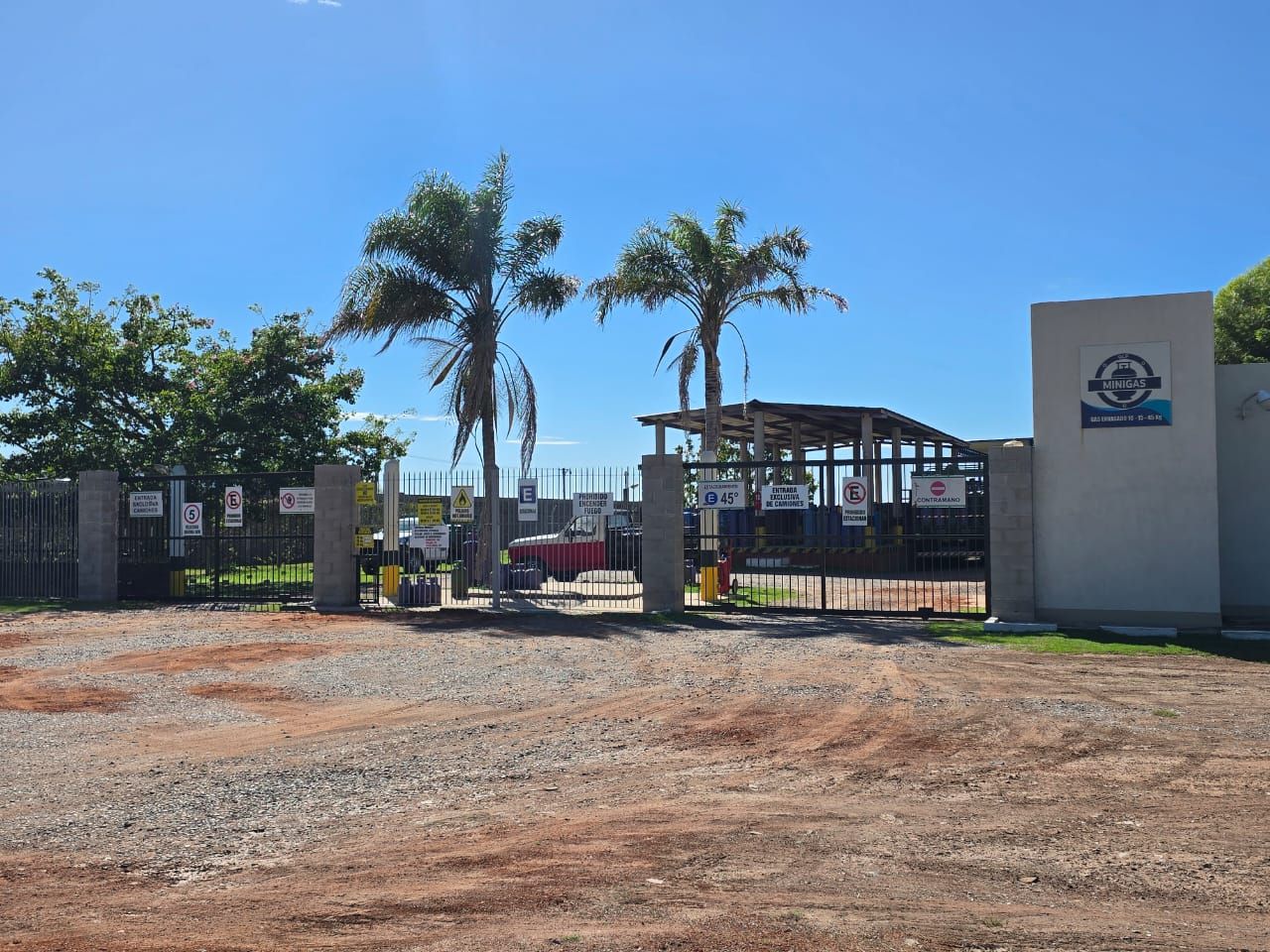 Un terreno de grava conduce a una entrada cerrada con dos palmeras y un muro de hormigón con un logotipo circular azul.