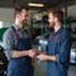 Two mechanics shaking hands in a garage; one hands keys to the other, smiling.