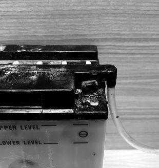 Black and white close-up of a damaged battery with a clear tube attached, showing signs of corrosion and leaks.