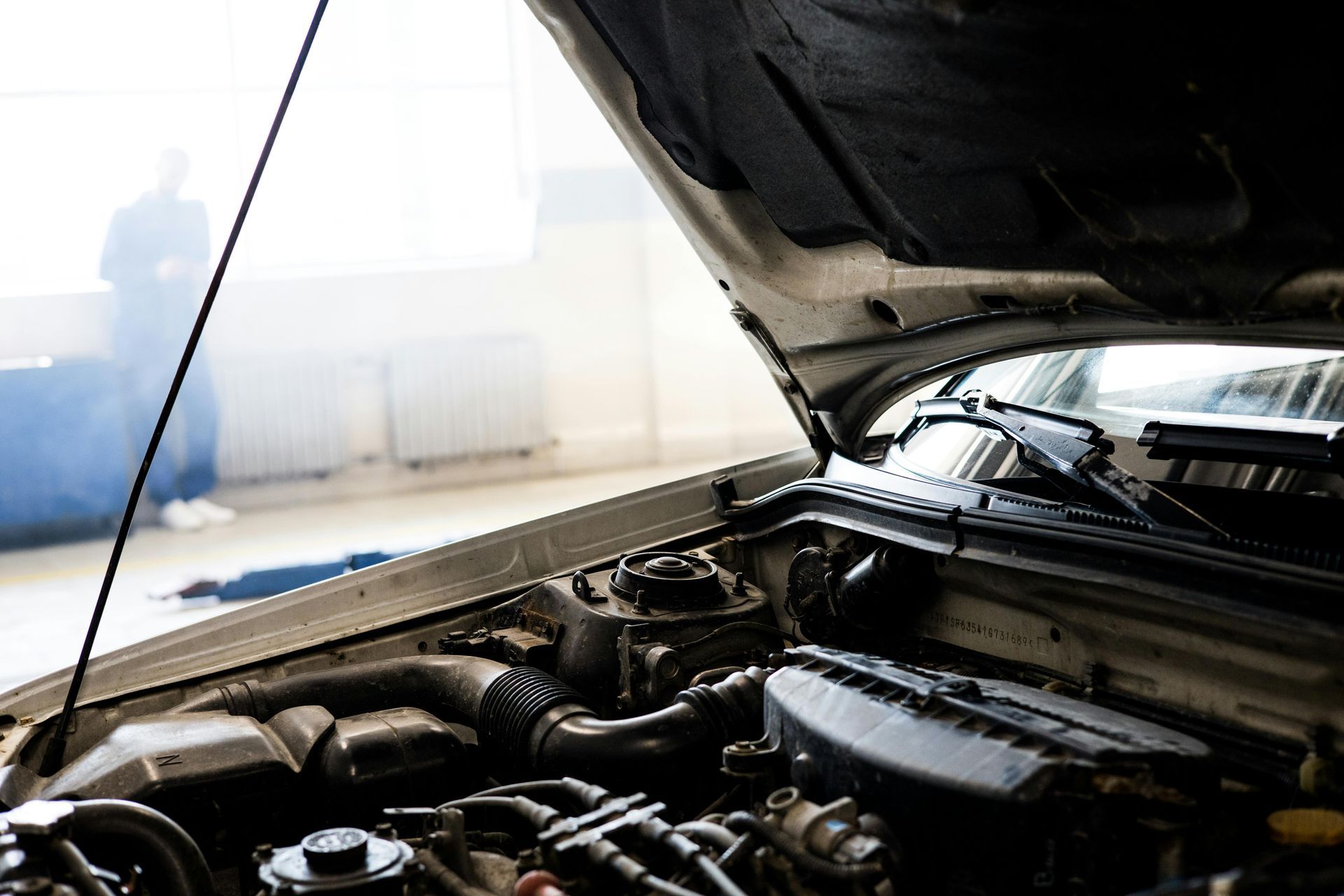 Car engine bay with the hood open, smoke rising; mechanic in the background.