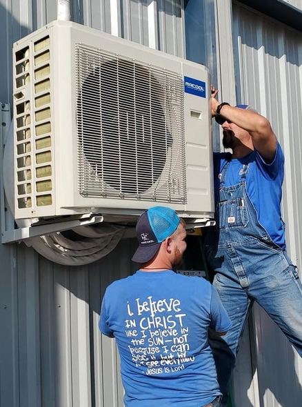 Two men installing an air conditioner unit on a corrugated metal wall.