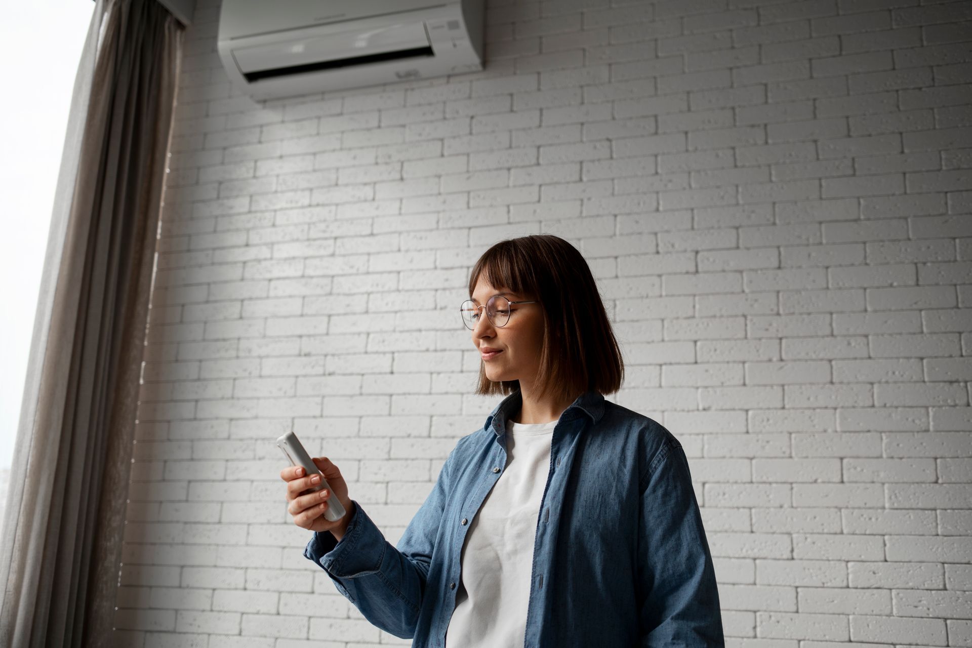 Woman operating an air conditioner remote control in a room with a white brick wall.