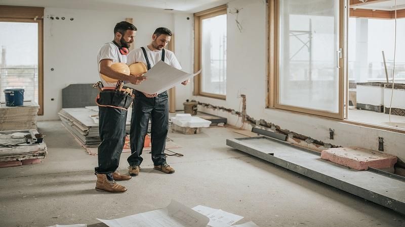 Two construction workers are looking at a blueprint in a room under construction.