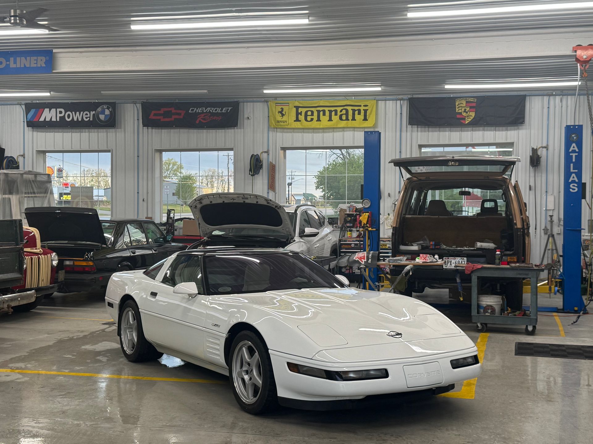 White Corvette in a garage with car flags; other vehicles in background.