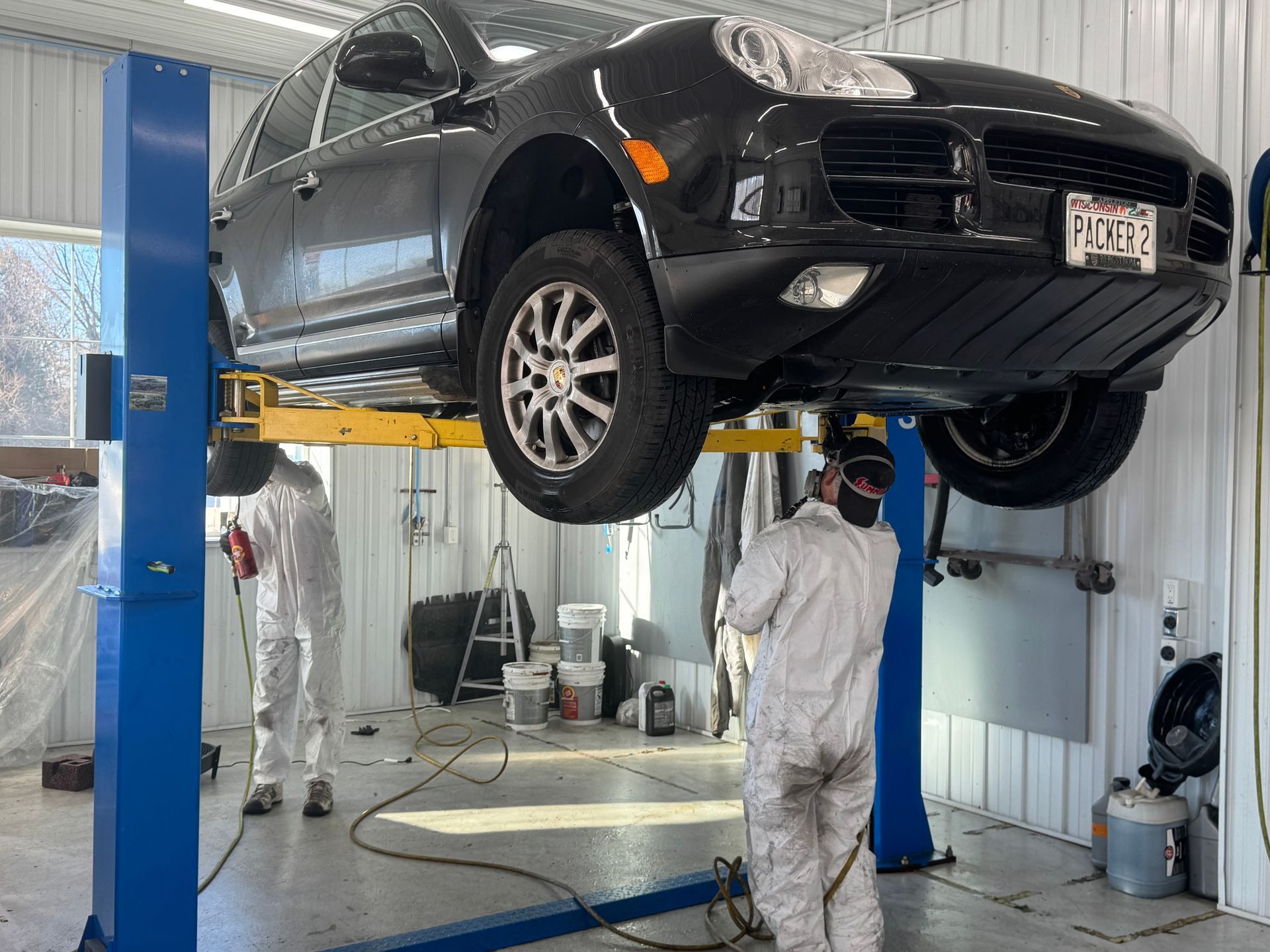 Two mechanics in white suits working on a black SUV raised on a lift in a garage.