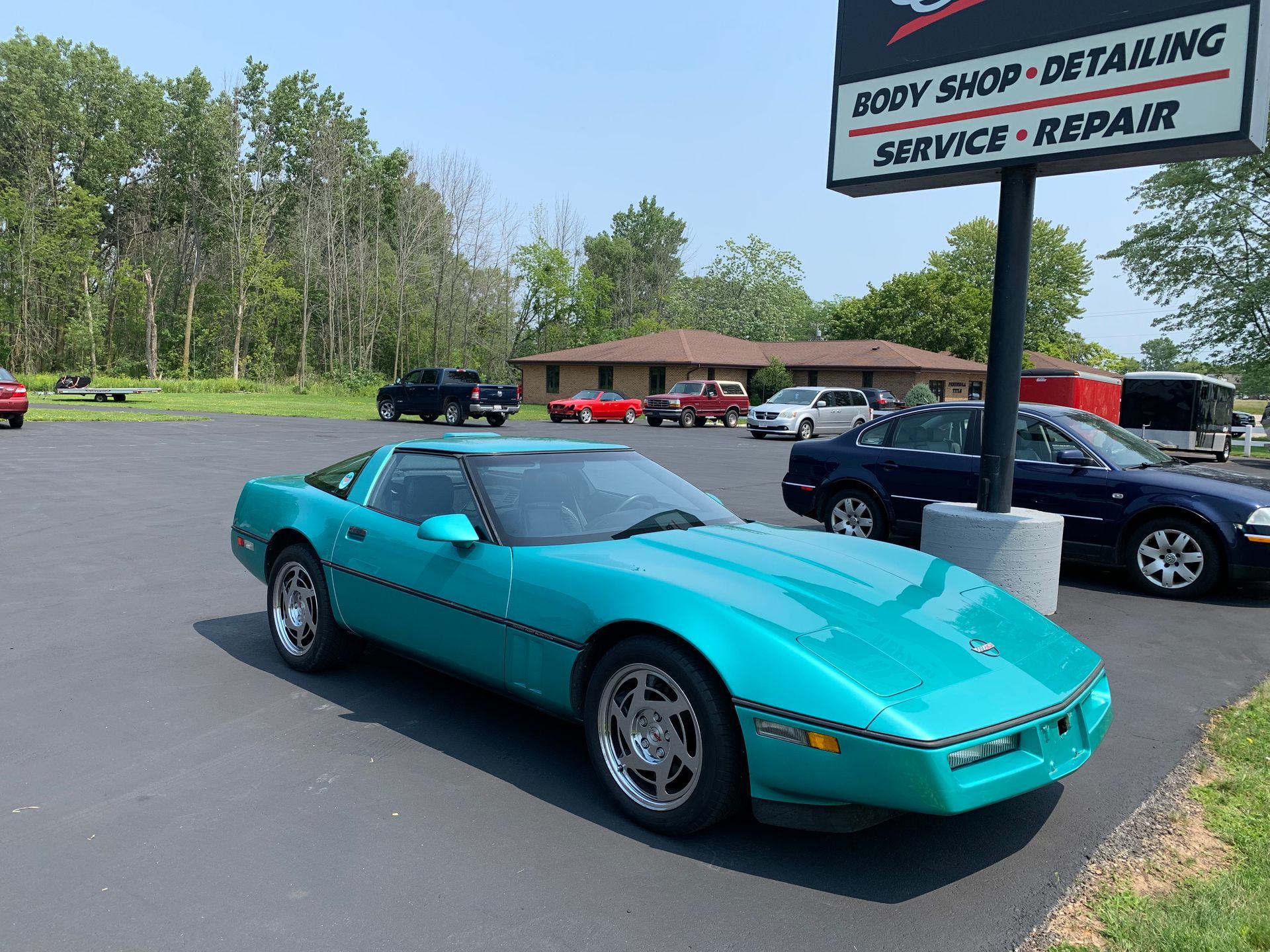 Turquoise Corvette parked in front of a body shop with other vehicles on a sunny day.