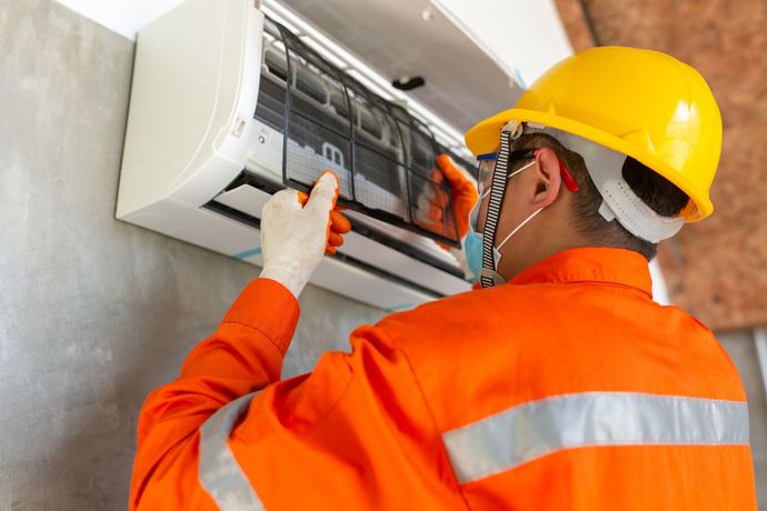 A man wearing a hard hat and mask is working on an air conditioner.