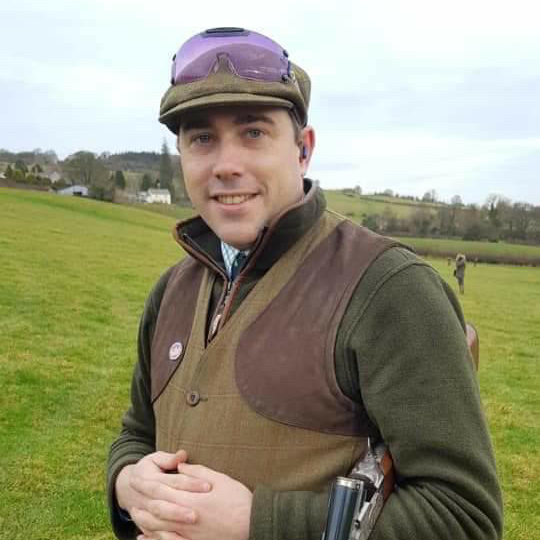 Person in a brown vest and green sweater, wearing a purple cap, standing on a grassy field holding a shotgun.