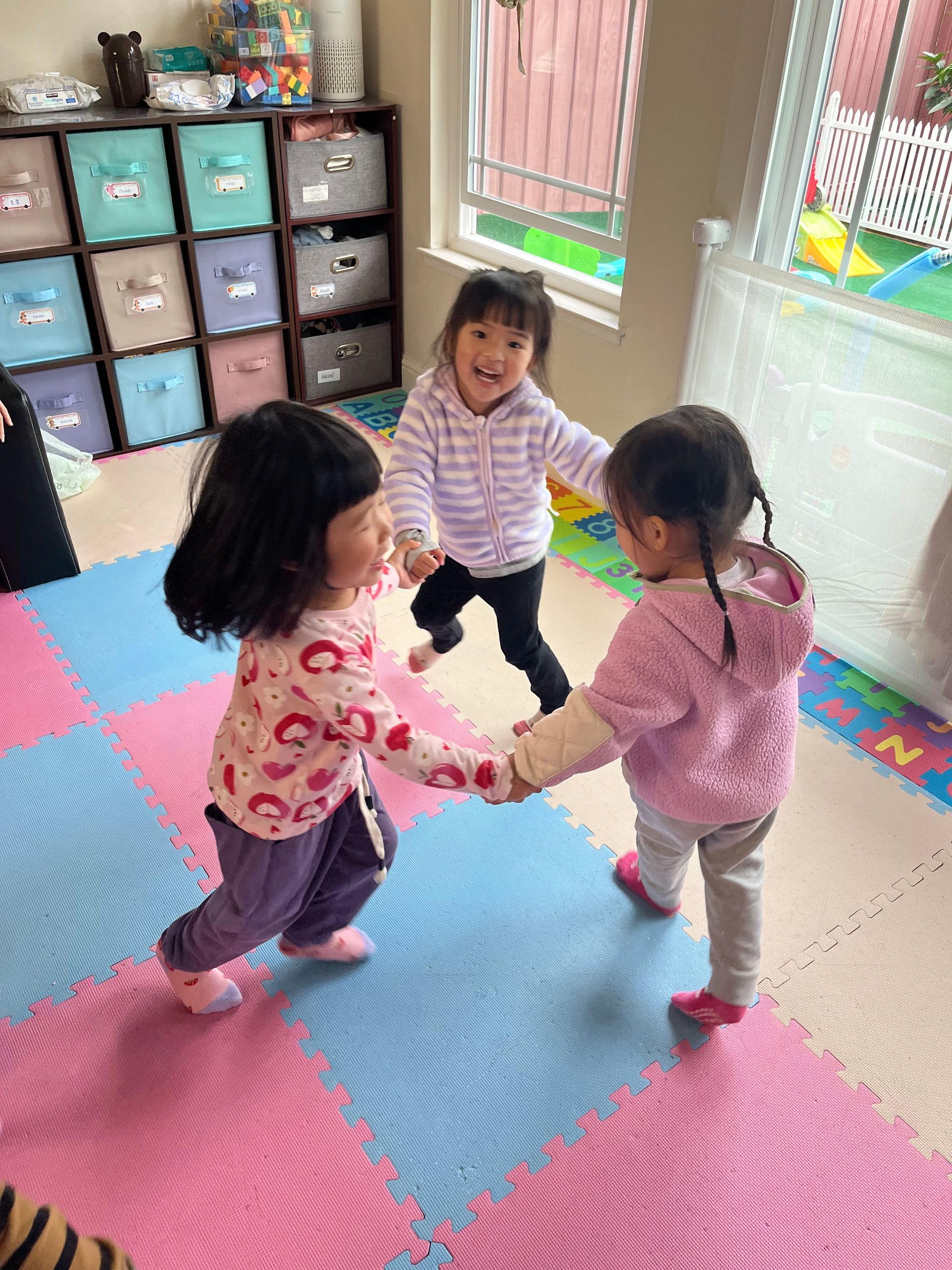 Three young children holding hands and playing on a colorful mat. Two wear pink, one wears purple and a striped sweater.