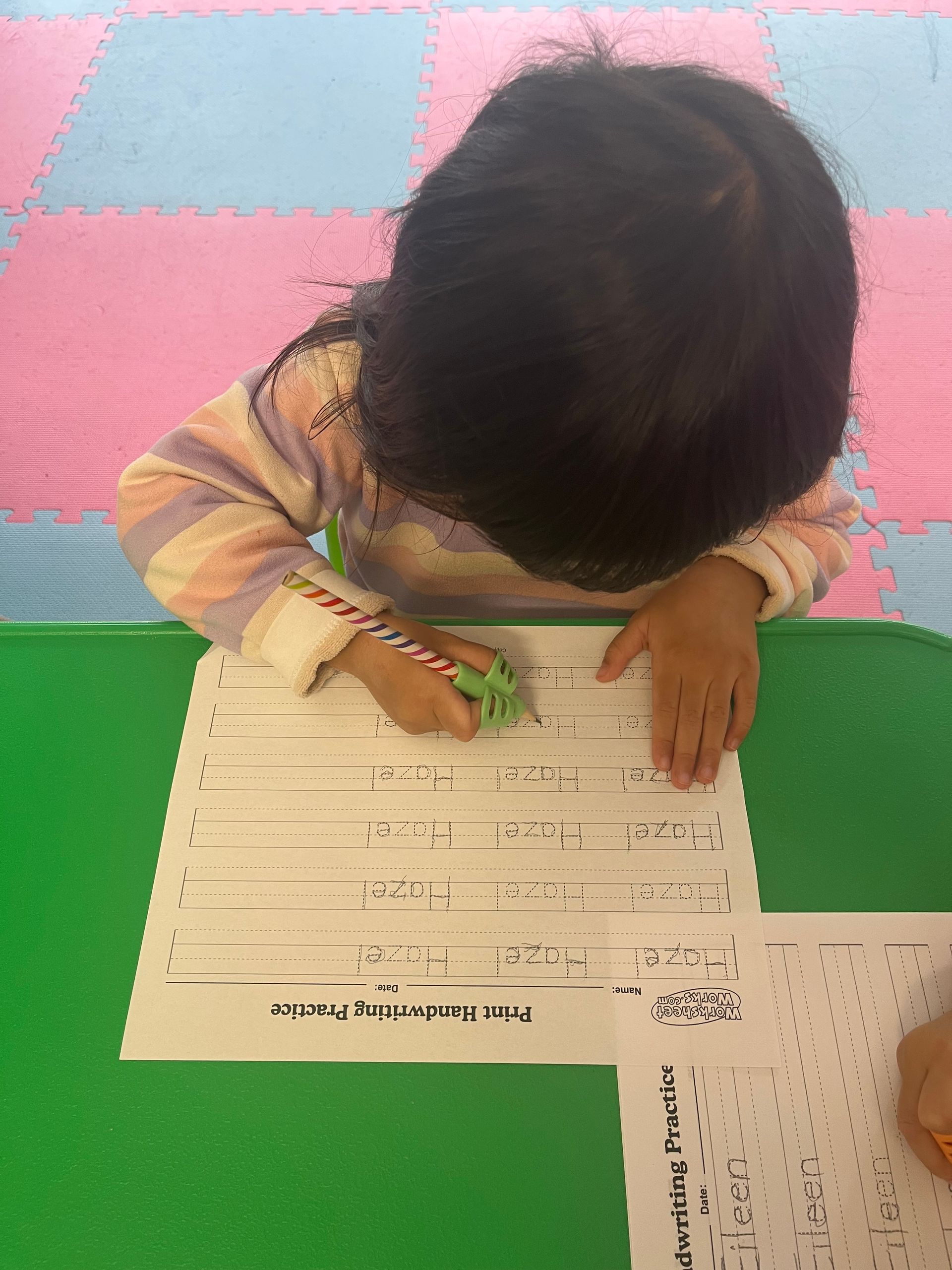 Child writing with a pencil on lined paper at a green table, pink and blue floor in the background.