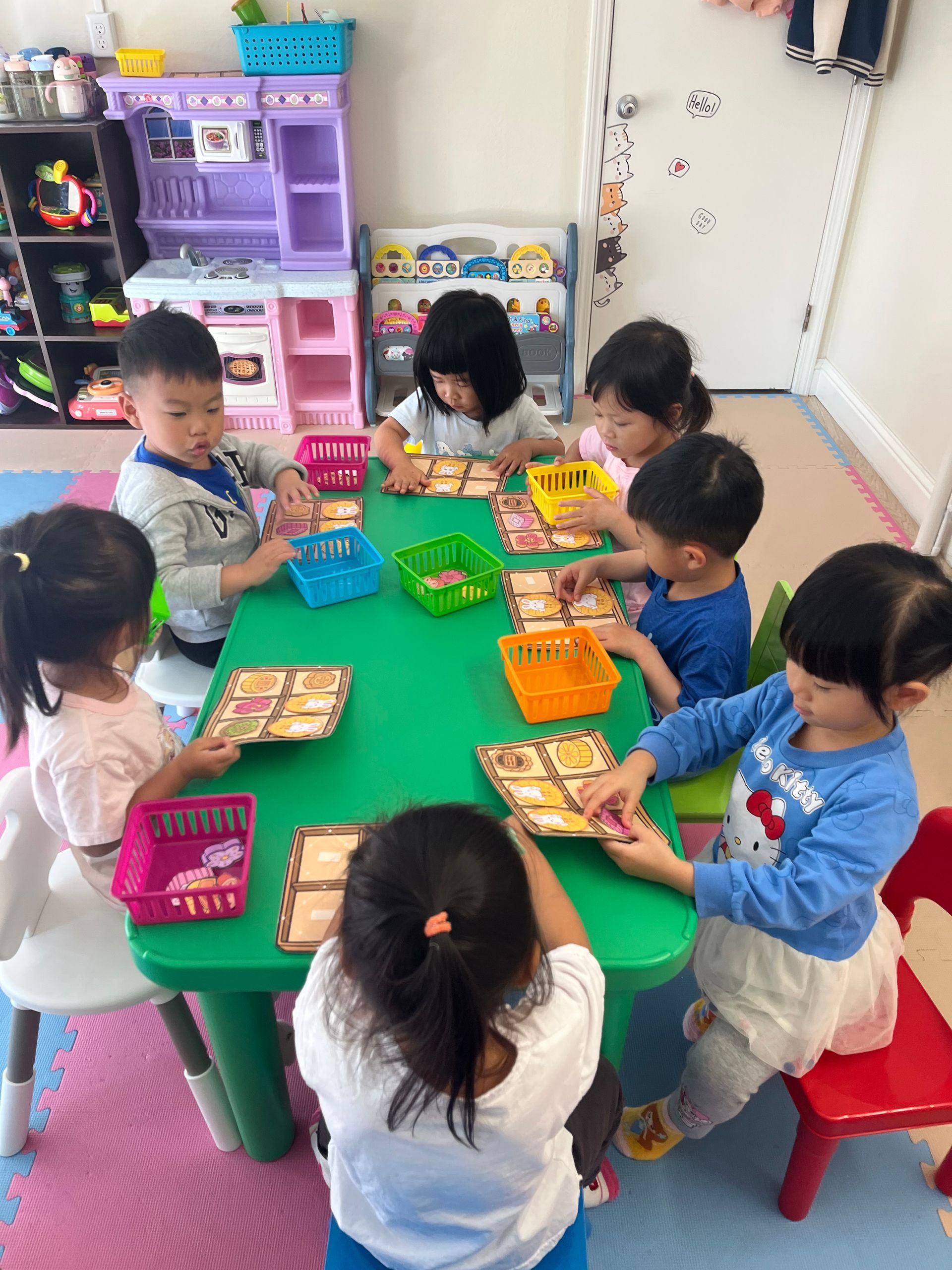 Children at a green table, each with a basket and card, playing a game in a colorful room.