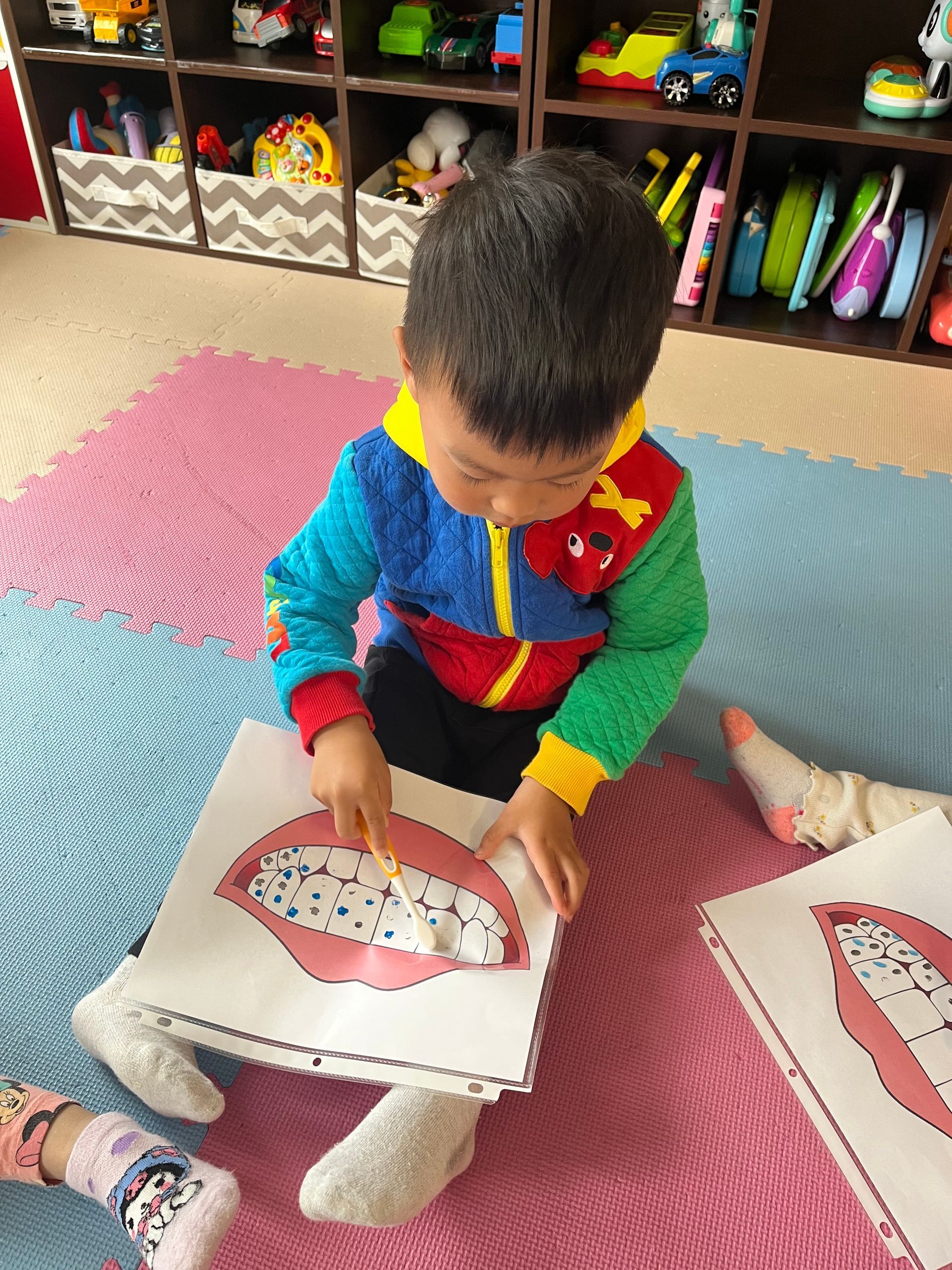 Child in colorful sweater looking at a paper cutout with white beads on pink mat in playroom.