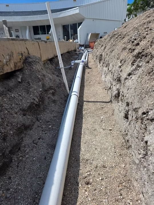 Trenched utility pipe installation beside a building, with gravel and dirt walls on both sides.