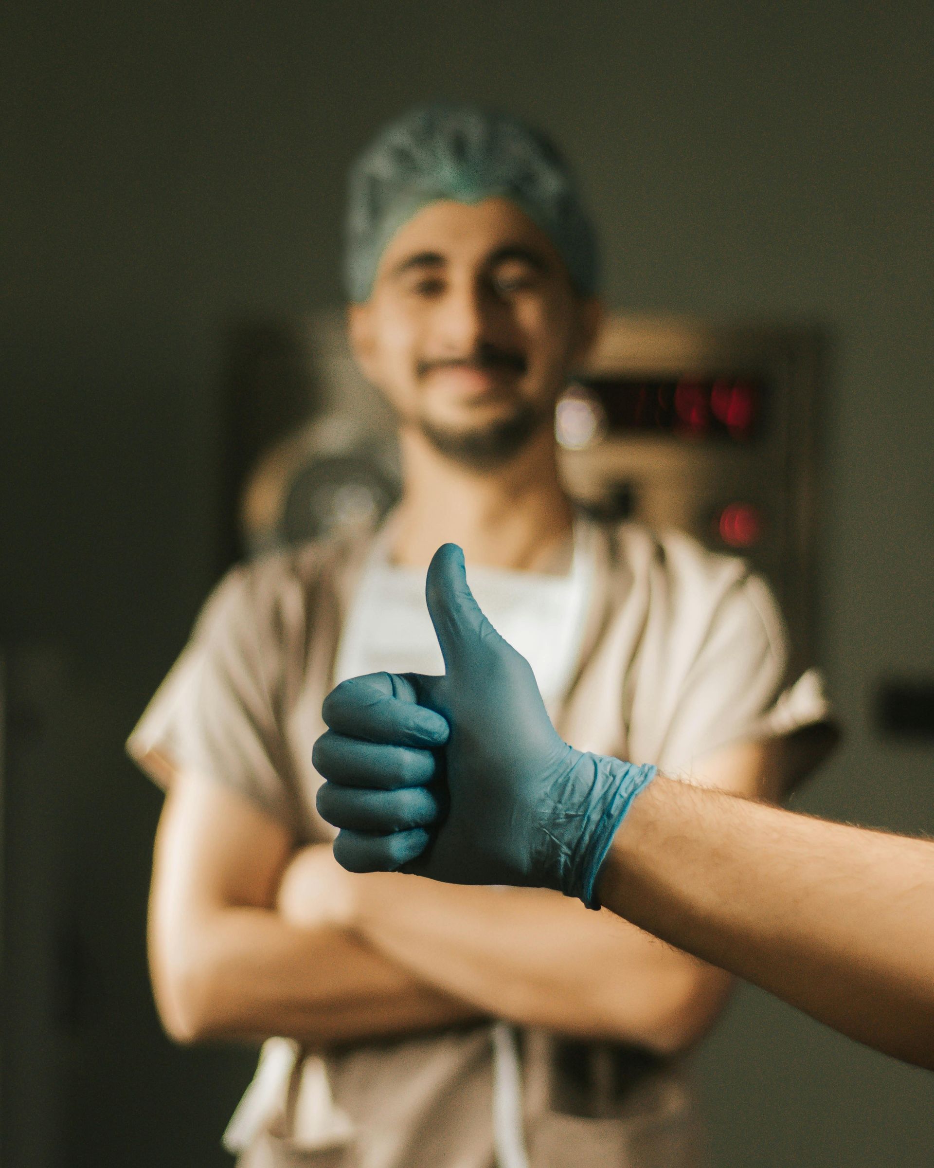 Gloved hand giving thumbs up in foreground, with a smiling doctor in surgical cap and scrubs behind.