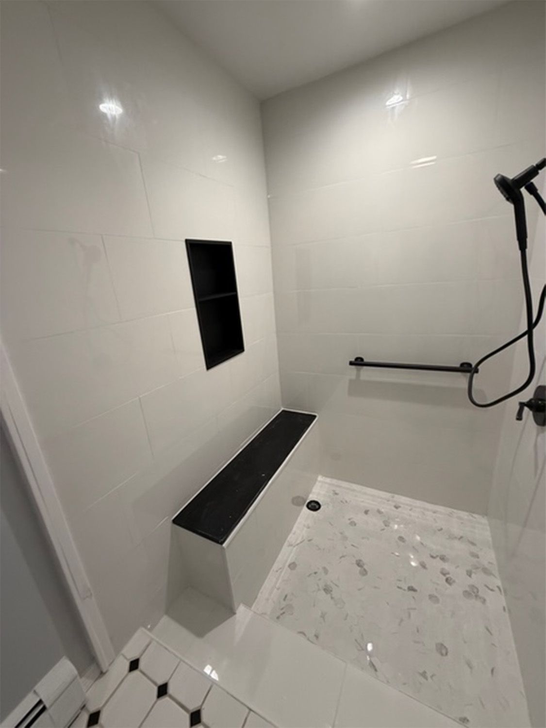 White tiled shower with black accents, built-in bench, grab bar, and recessed shelf. Marble-look floor.
