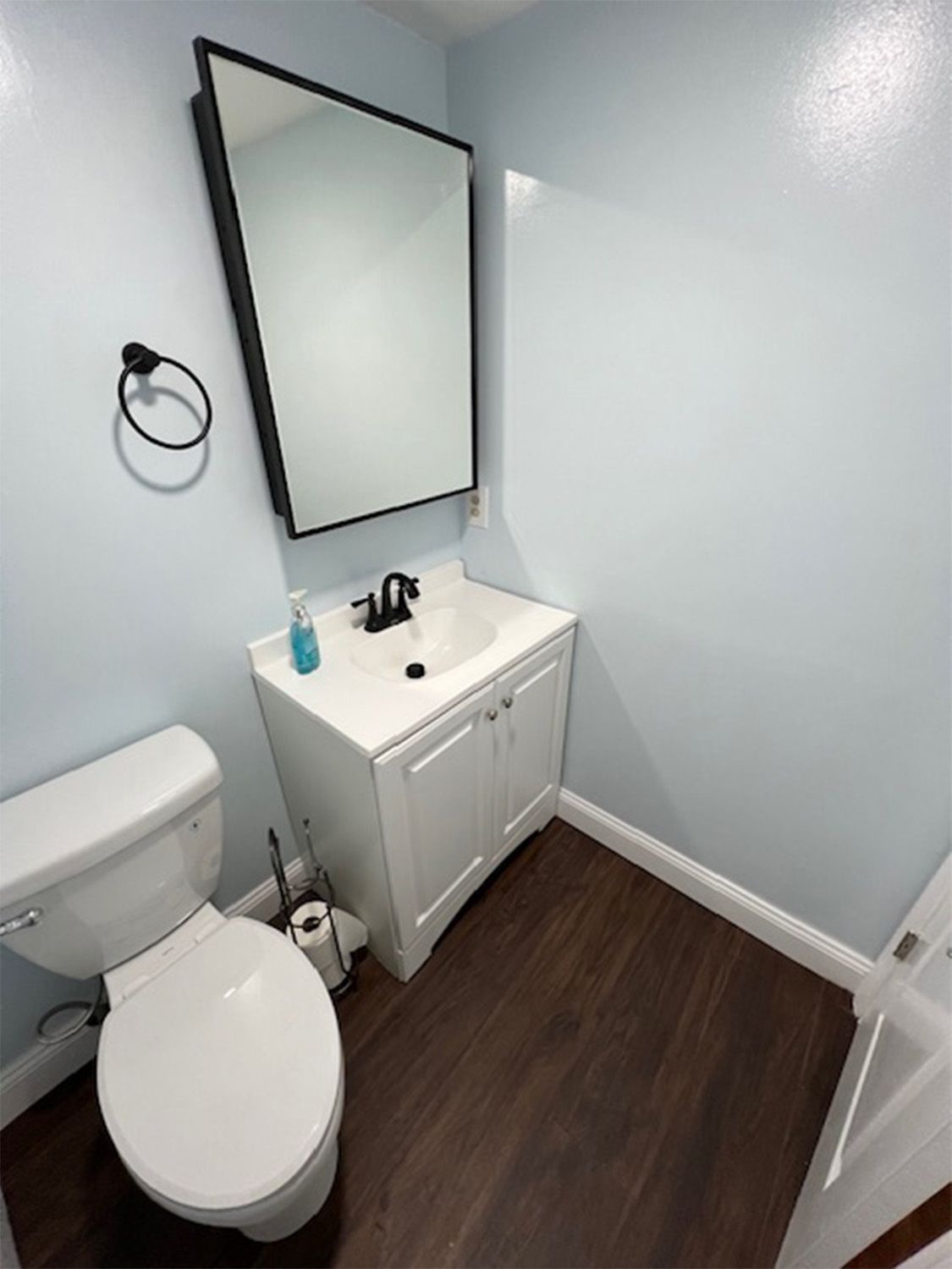 Small bathroom with a toilet, sink, and mirror; light blue walls, dark wood floor.