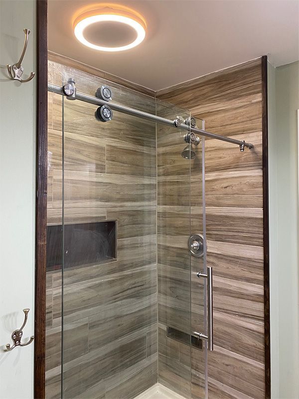 Shower with sliding glass door, brown wood-look tile, stainless steel hardware, and a round ceiling light.