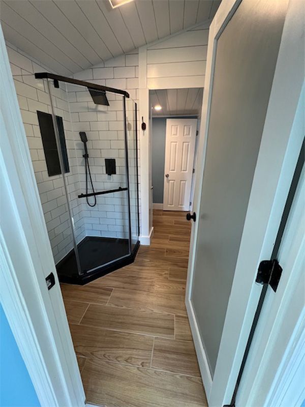Bathroom with shower, wood-look floor, and white subway tile walls. Doorway leads to a hallway with a closed door.