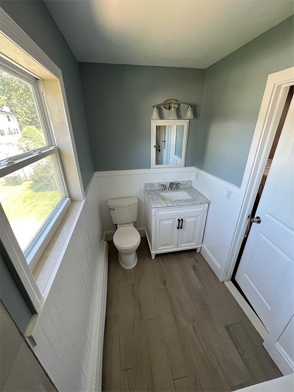 Bathroom with white wainscoting, teal walls, vanity, toilet, window, and wood-look flooring.