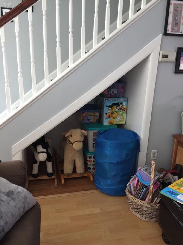 Under-stair storage area with toys, rocking horses, and blue laundry hamper. Light wood floor, white and blue walls.