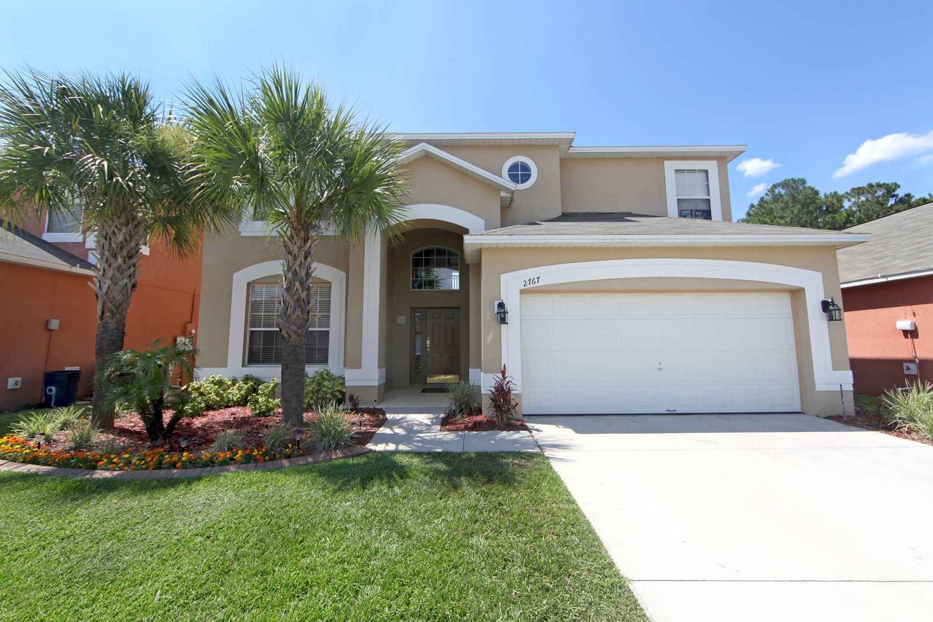 Two-story beige suburban house with palm trees, white garage, and a bright green lawn