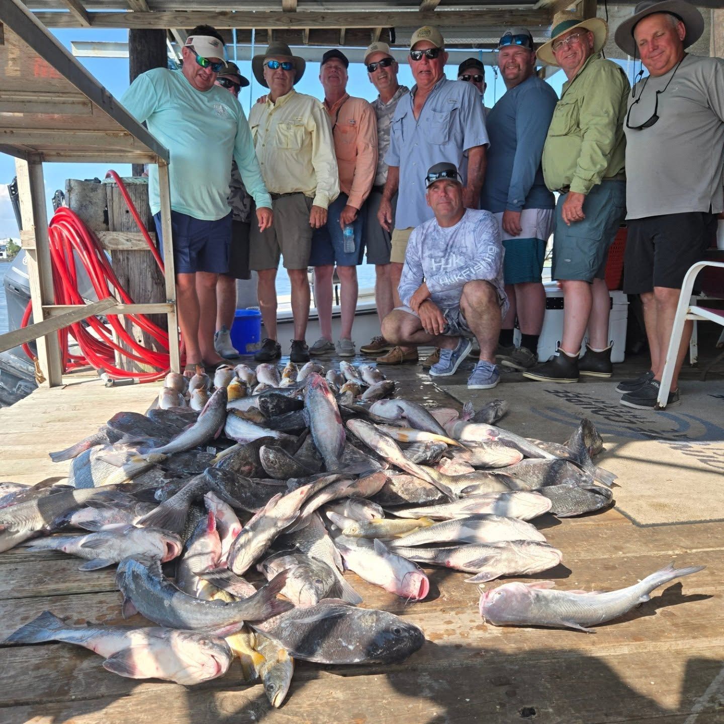 Group of people standing behind a large pile of fish on a dock, with nets and fishing gear nearby.