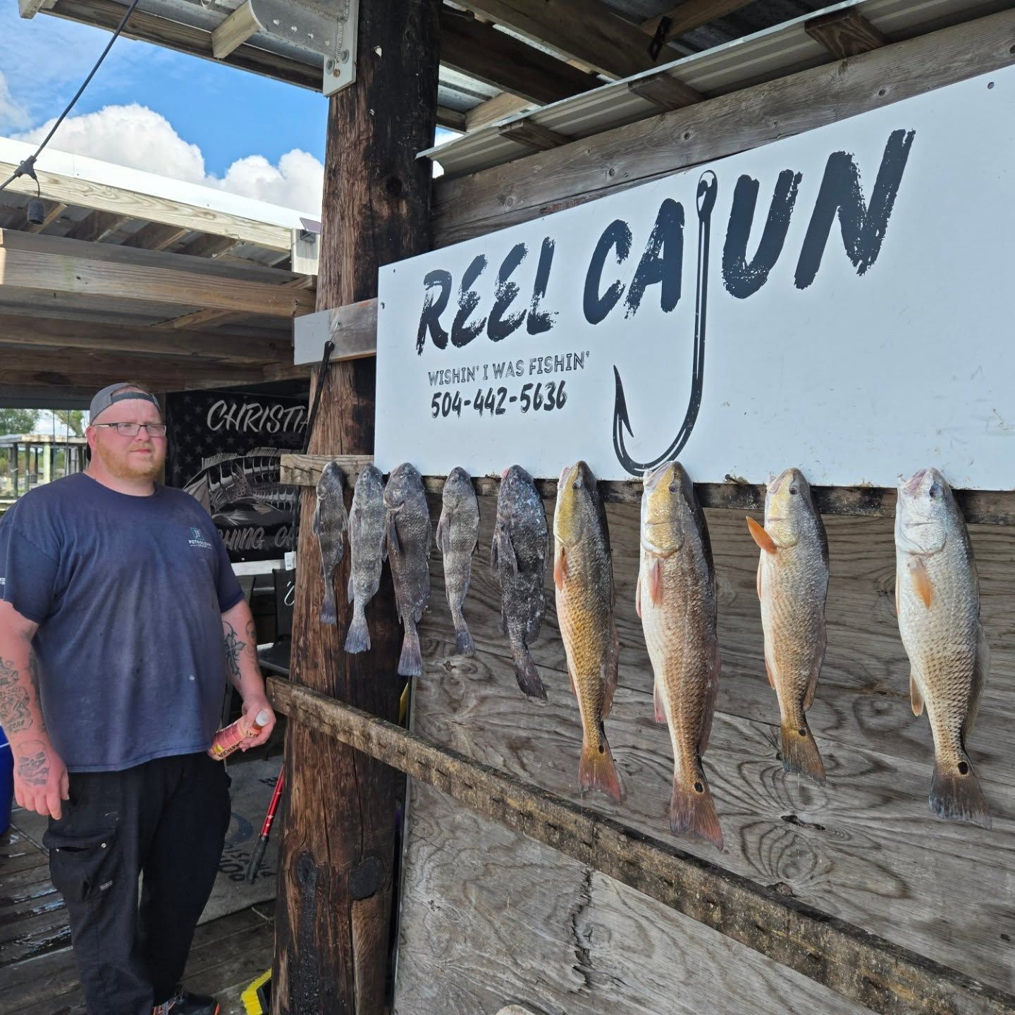 Man standing beside a sign at Reel Cajun, with several fish hanging on a dock wall.
