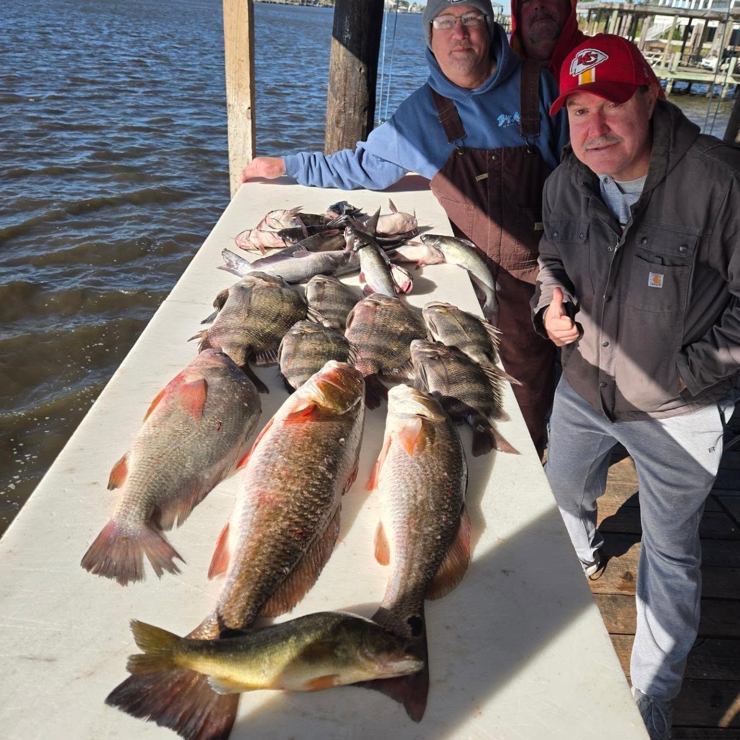 Two people on a dock display several large fish beside the water.