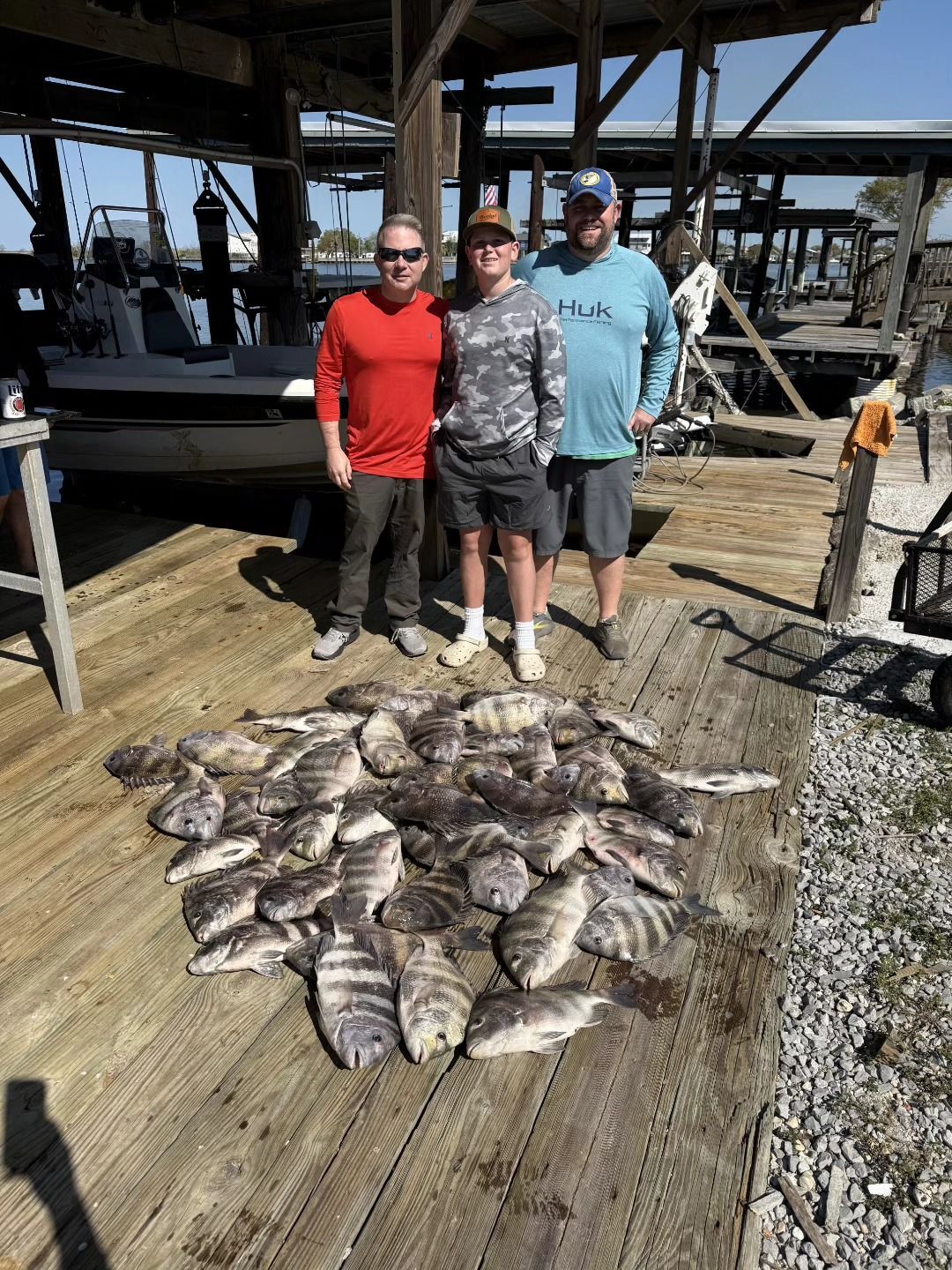 Three anglers pose by a dock with a large pile of freshly caught fish.