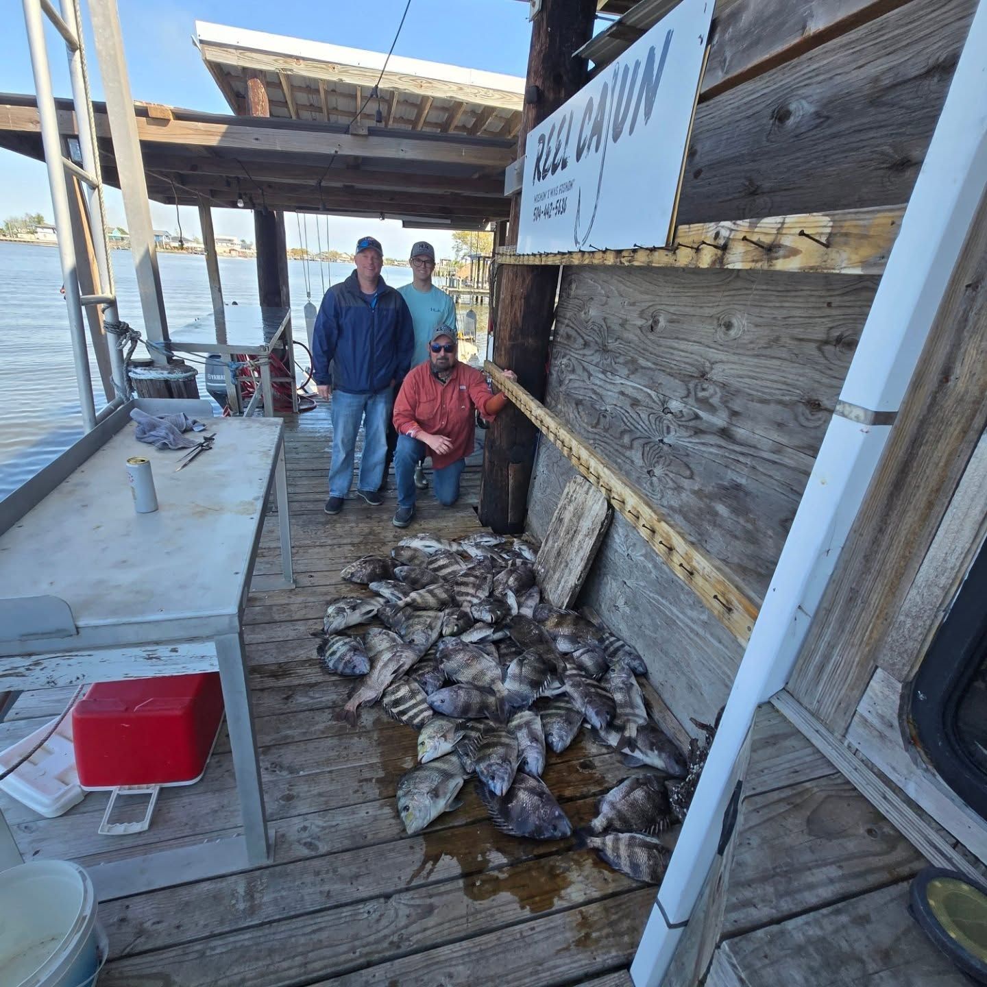 Two people stand by a pile of fish on a wooden dock beside the water.