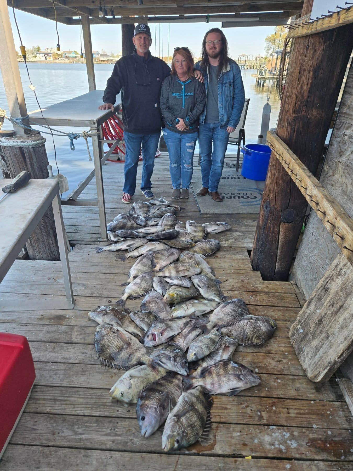 Three people standing on a dock beside a large pile of fish, with water and boats in the background.