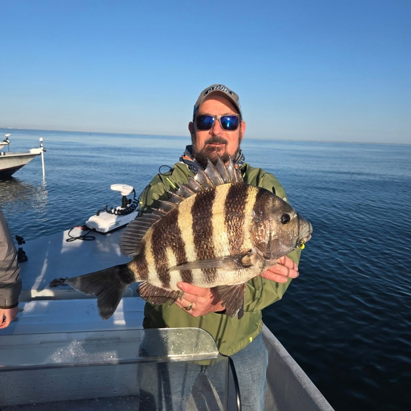 Man holding a large striped fish on a boat, with calm water and clear blue sky in the background.