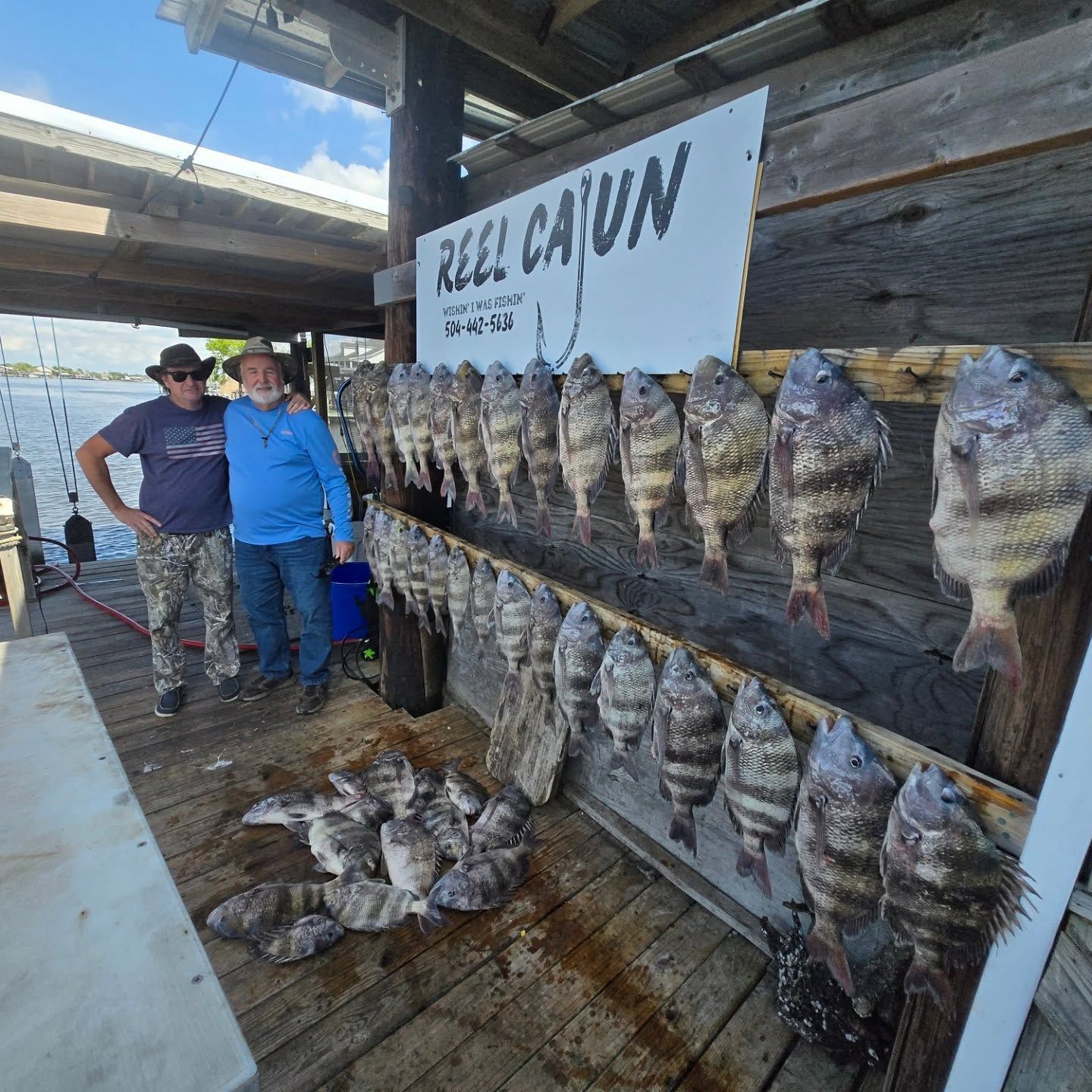 Two people beside a wall of hanging fish at a rustic seafood stall under a covered dock.