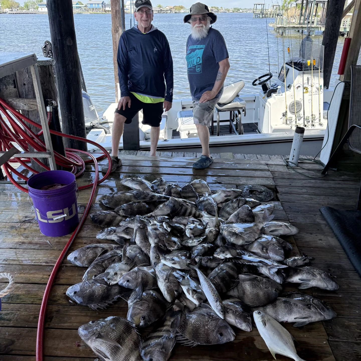 Two people stand by a large pile of fish on a dock, with boats and water in the background.