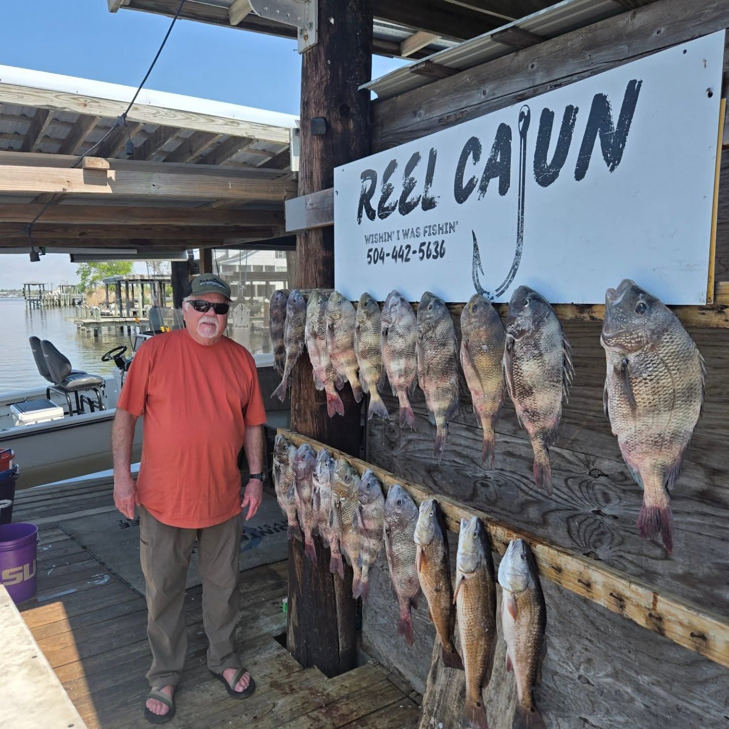 Man standing beside a dockside fish display with rows of hanging fish and a “REEL CAJUN” sign