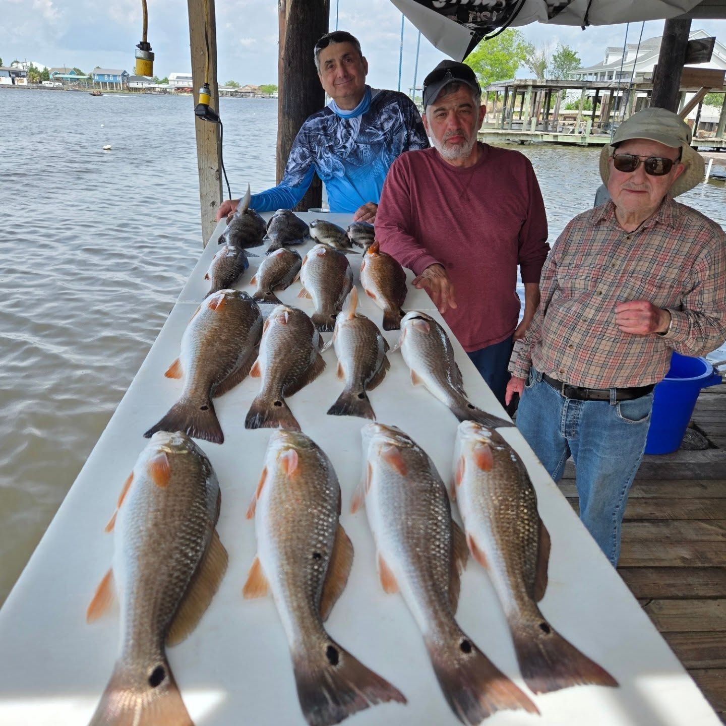 Three people pose beside a table of freshly caught fish on a dock by the water.