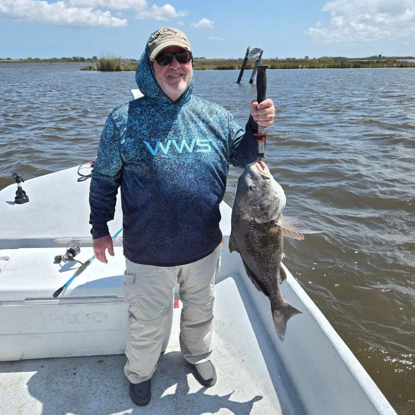 Angler on a boat holding a large fish, wearing a blue hoodie and cap on a cloudy day.