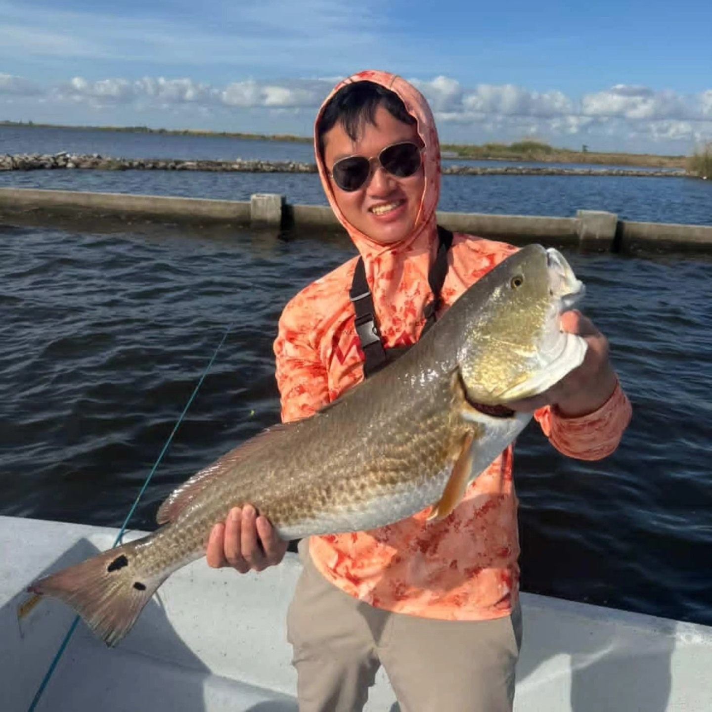 Person on a boat holding a large fish, wearing sunglasses and a hooded orange shirt.