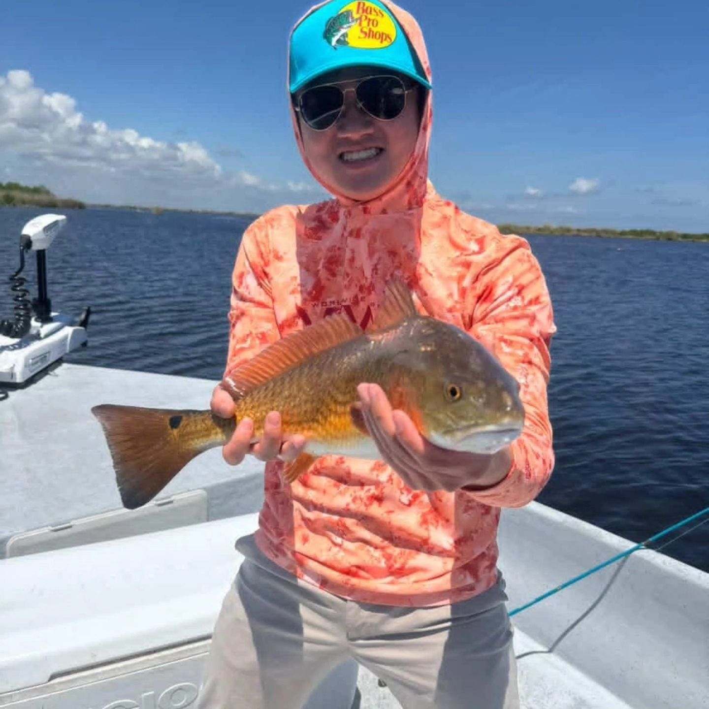 Person on a boat holding a large fish, wearing a teal cap and coral shirt on sunny water.