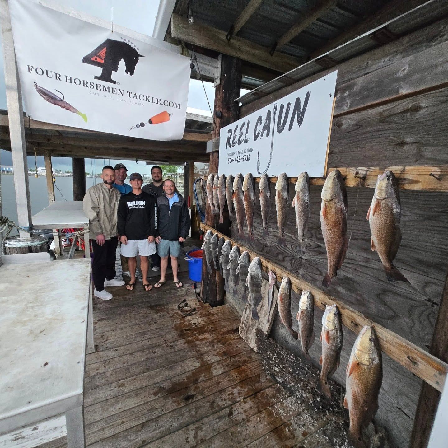 People posing beside a wall of hanging fish at a rustic dockside fish cleaning area.