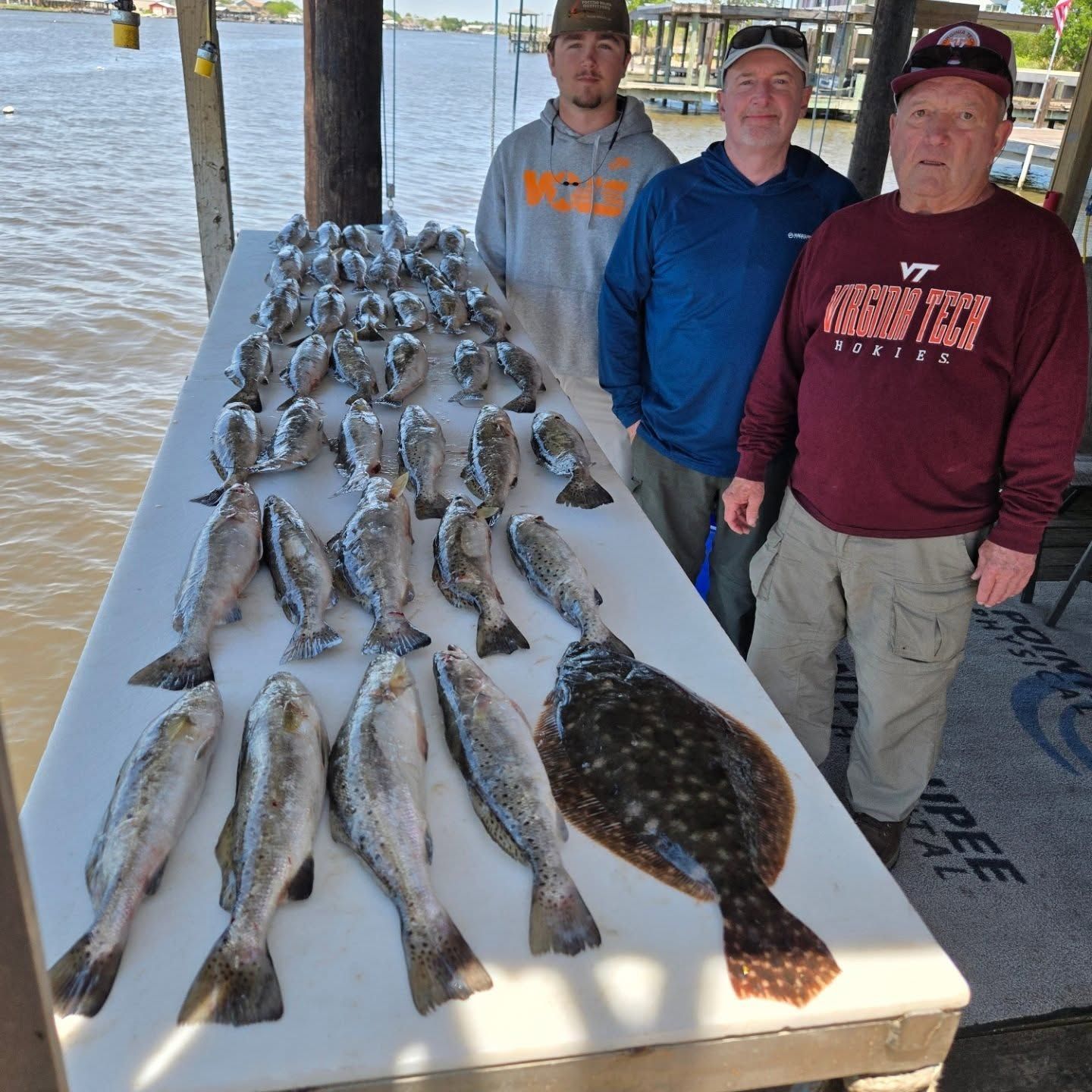 Three people stand beside a table covered with fish on a dock by the water.