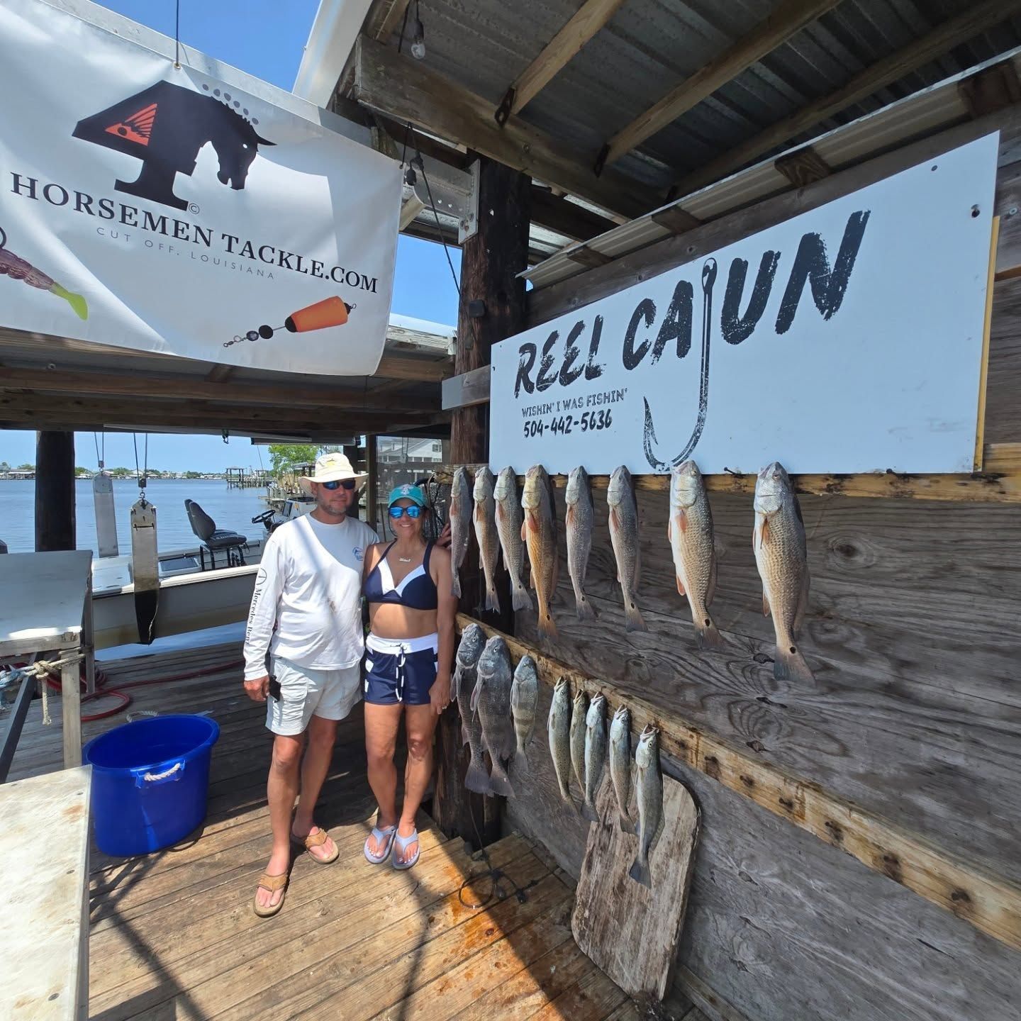 Two people posing beside hanging fish at a rustic dock seafood shack under “BULL CAJUN” signs