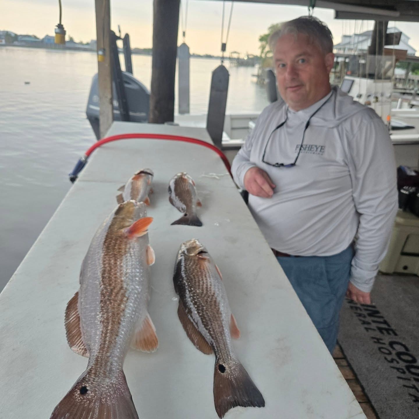 Man on a boat beside three fish laid on a table at sunset with water in the background