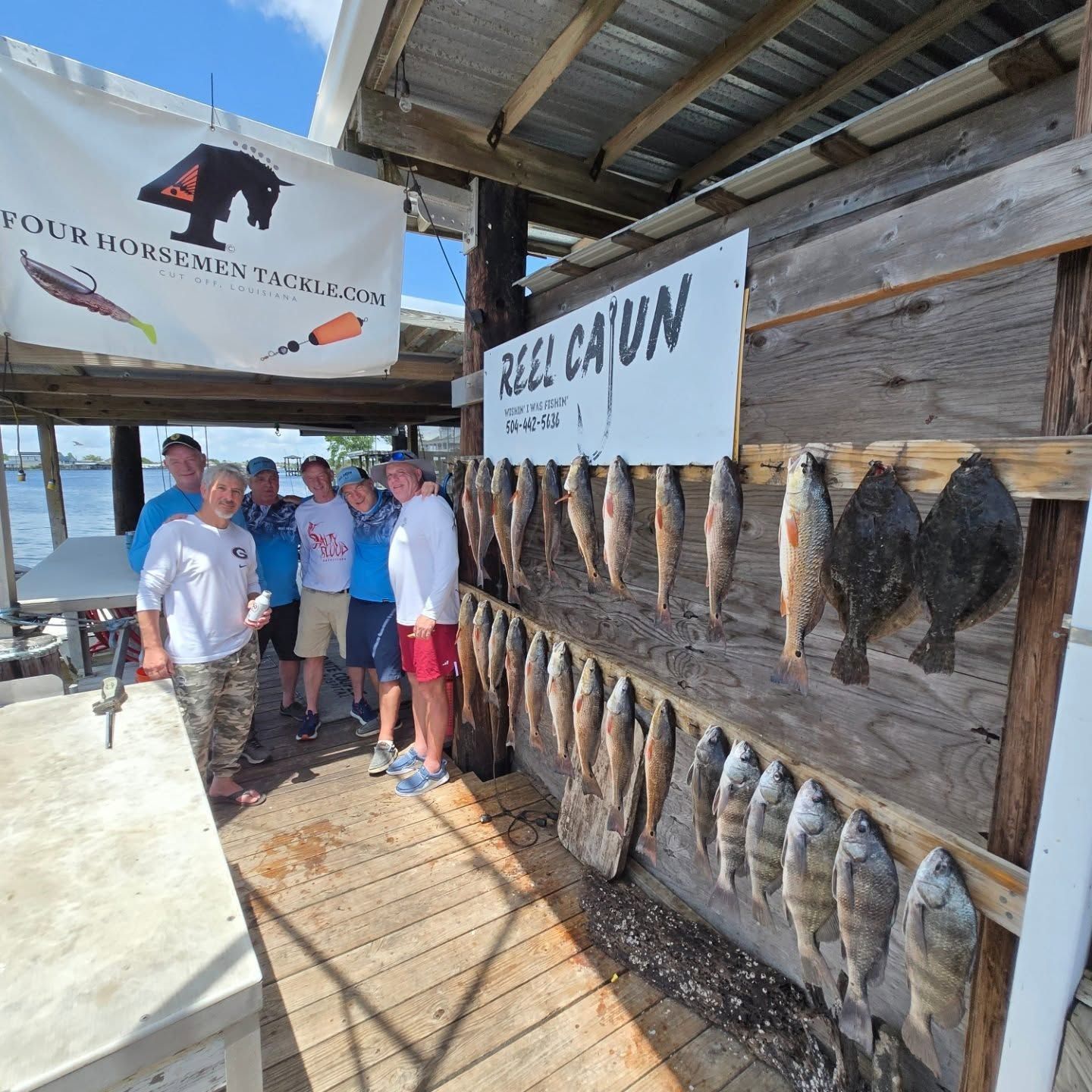 People posing beside a “Hill Cajun” fish display at a rustic seafood shack outdoors