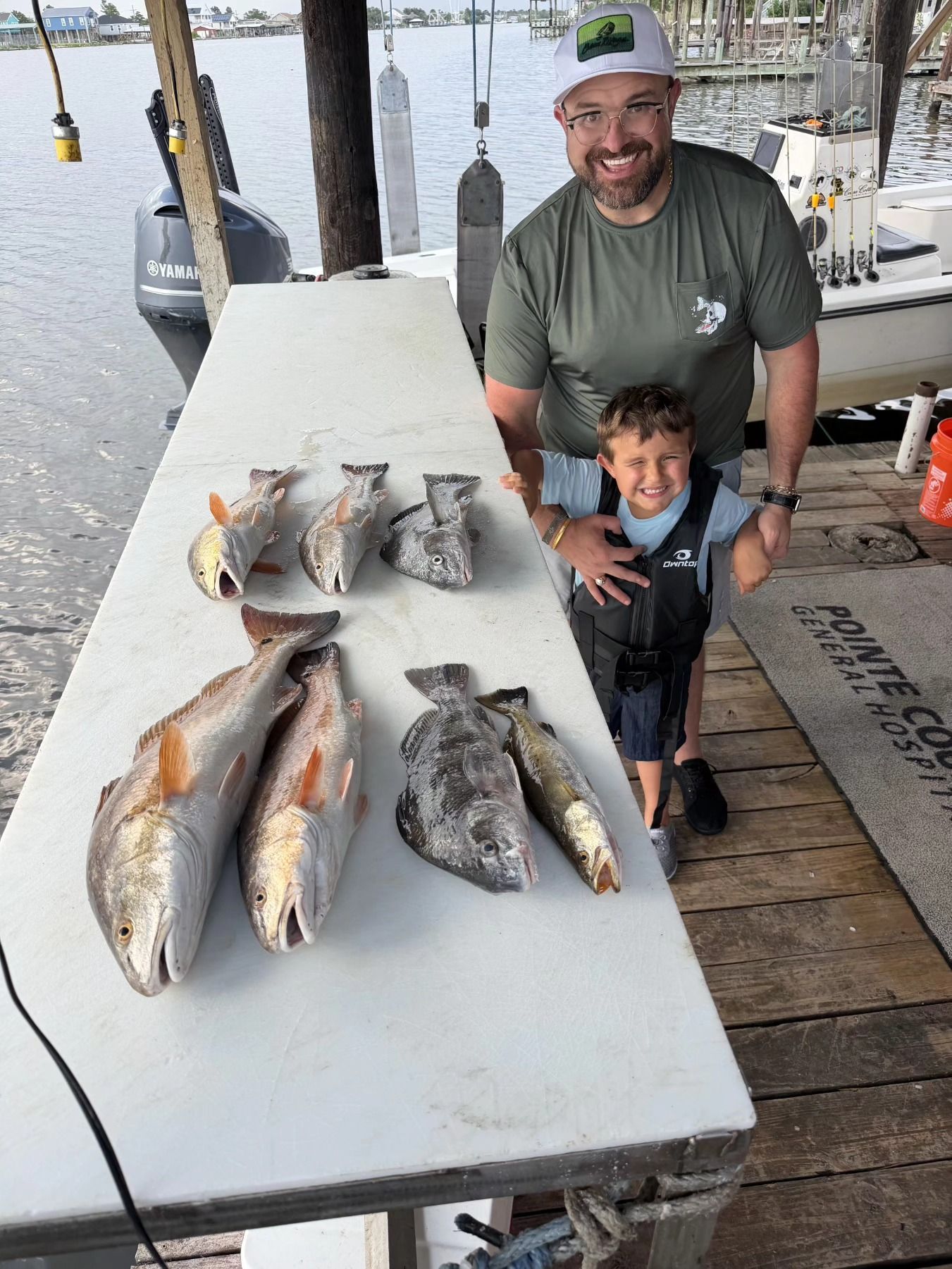 Man and child posing on a dock beside several large fish laid out on a table.