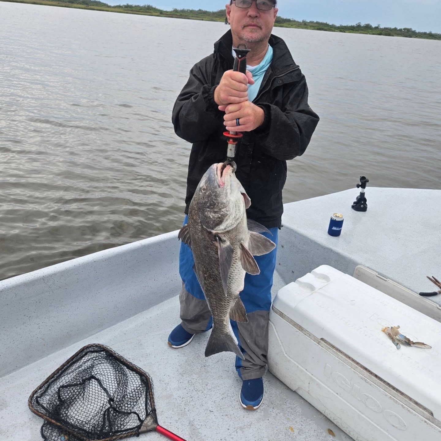 Person on a boat holding a large fish over calm water, with a net and cooler nearby.